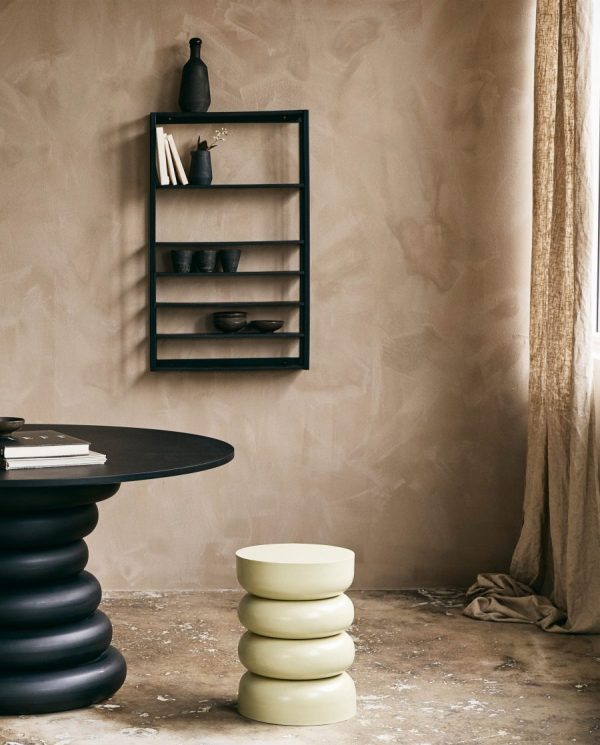 Et minimalistisk værelse med teksturerede beige vægge har en NORDAL - Zaros Hylde med forskellige mørke dekorative genstande. ved siden af er der et unikt sort cirkulært bord og en stak af tre grønne afrundede taburetter.