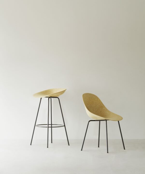 To moderne stole mod en almindelig væg; den ene er en høj skammel med sort metalstel og rundt korksæde, den anden en standardhøjdestol med matchende stel og et ergonomisk formet korksæde - NORMANN COPENHAGEN - Mat Spisebordsstol.
