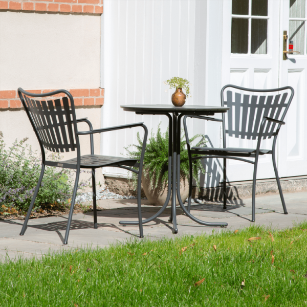 To sorte metalstole og et rundt bord sat på en terrasse, med designmøbler-æstetik, med en lille potteplante på bordet. Baggrunden viser en hvid dør og en del af et murstenshus, indrammet af grønt græs.