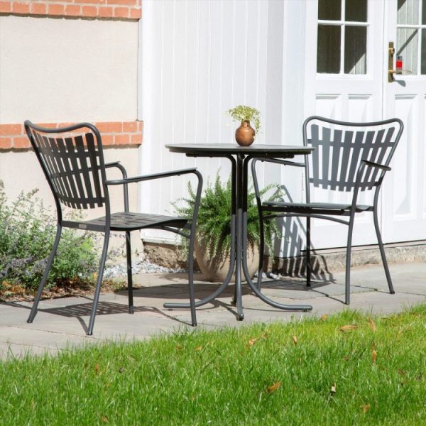 En lille terrasse udenfor et hus med to sorte metalstole og et matchende rundt bord. En lille plante i en vase står på bordet, alt sammen sat på baggrund af en hvid dør og vinduer, og fremviser udsøgt møblerdesign.