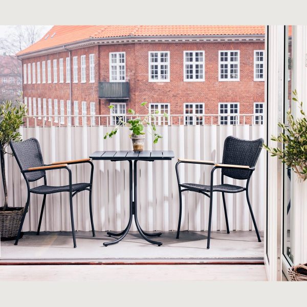 En hyggelig altan med to sorte kurvestole og et lille rundt bord, der viser en potteplante. Balkonen har udsigt over en klassisk murstensbygning og er omkranset af et hvidt træhegn, der afspejler det seneste inden for TomorrowsDesign-æstetik.