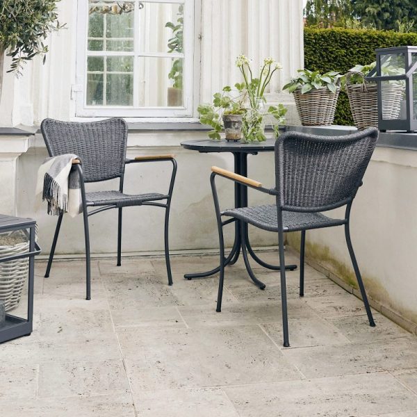 To moderne sorte kurvestole med træarmlæn og designmøbler ved siden af et lille rundt bord på en flisebelagt terrasse. Et gråt tæppe er draperet over den ene stol, og potteplanter er synlige i baggrunden.