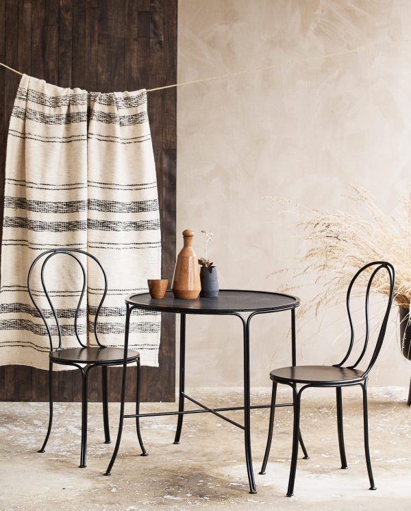En rustik spiseplads med et sort metalbord og to matchende stole, en beige væg, et træpanel og et hængende stribet klæde. TomorrowsDesign inkluderer en vase og træredskaber på bordet ved siden af tørrede planter.