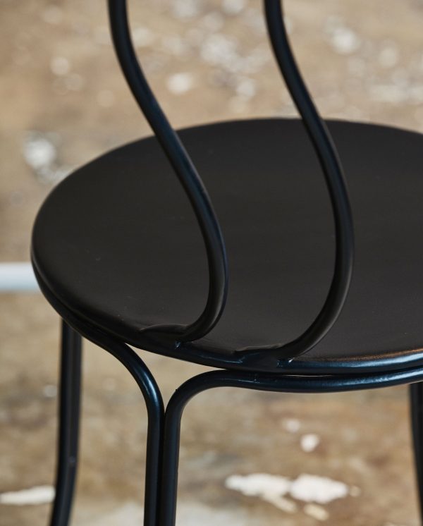 Nærbillede af en moderne sort metalstol med et cirkulært sæde og et elegant, looping ryglænsdesign, placeret mod en struktureret, neutral baggrund i et interiør.