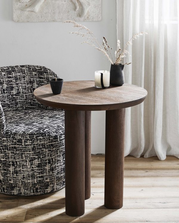 Et hyggeligt indvendigt hjørne med et lille rundt træbord med en mønstret stol ved siden af. På bordet står en sort vase med tørrede blomster og et lille lys. Lysgardiner designet af TomorrowsDesign hænger i baggrunden.