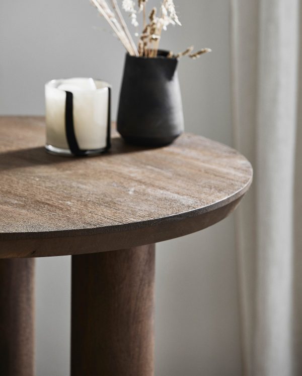 Et nærbillede af et rundt træbord med en sort vase med tørrede planter og et hvidt lys, mod en blød grå baggrund med designmøbler gardin.