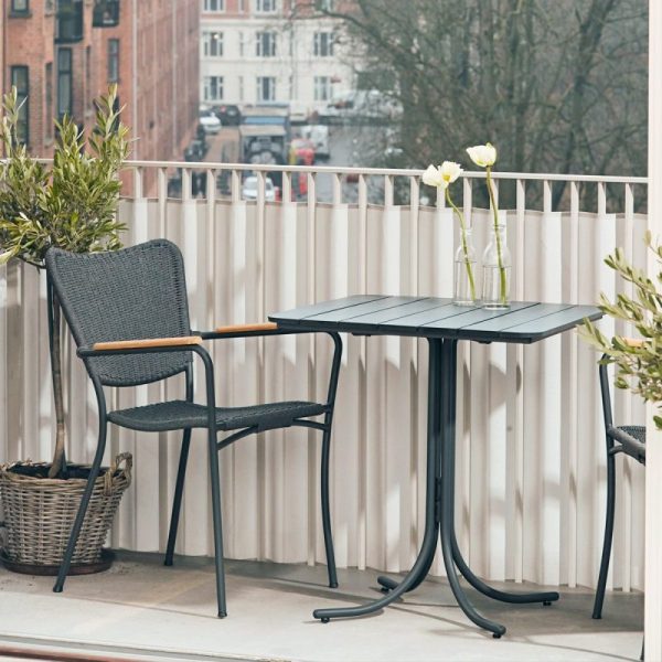 En lille altanopstilling med en mørk kurvestol og et matchende mørkt bord, som har en hvid blomst i en vase og en blyant. Der er potteplanter og en vævet kurv ved siden af bordet med udsigt over en gade. Møblerne fremhæver rummets interiør-æstetik.