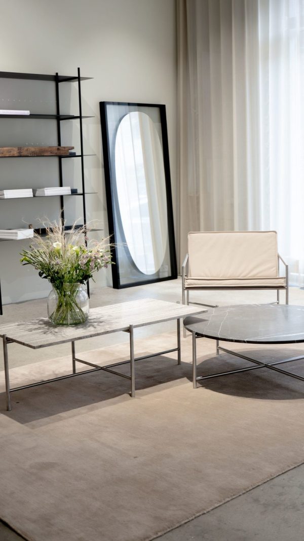 En moderne stue med en minimalistisk stol og sofabord på et beige tæppe, et højt spejl lænet op ad væggen, en reol og en vase med blomster på designmøblerbordet.