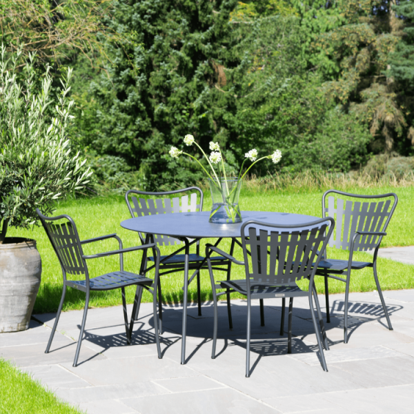 Et moderne udendørs spisesæt med et blåt bord og fire matchende stole har et elegant design. Opsætningen er på en terrasse omgivet af en frodig grøn have og træer. En vase med hvide blomster stilles på bordet.