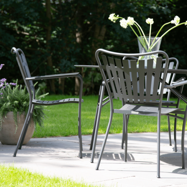 Et terrassesæt med fire grå stole omkring et matchende bord, designet af TomorrowsDesign, placeret på et brolagt område i en frodig have. En vase med hvide blomster står på bordet.