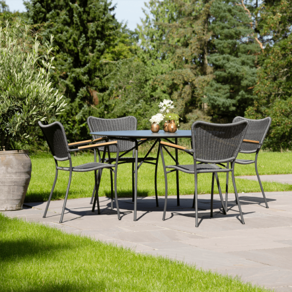 Et moderne udendørs spisesæt bestående af et metalbord og fire vævede stole på en terrasse, omgivet af frodig grøn græsplæne og en række planter under klar himmel, afspejler principperne i TomorrowsDesign.