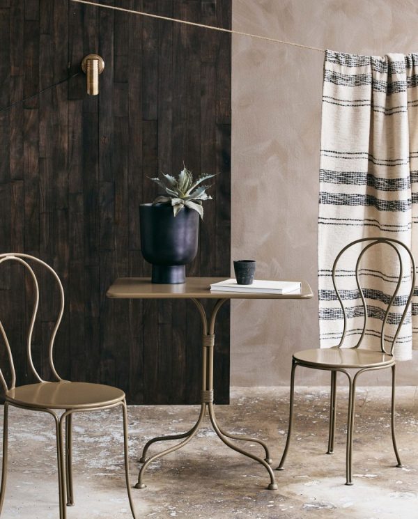 Et hyggehjørne med et NORDAL - Viola Havebord med to metalstole, en mørk vase med en plante og en kop. Et stribet gardin hænger i baggrunden, og indstillingen viser en delvis trævæg og tekstureret gulv.