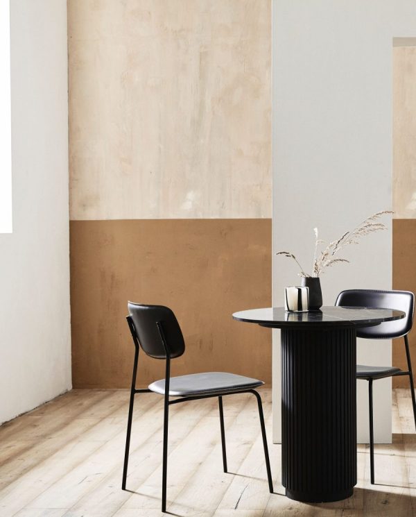 En minimalistisk spisestue med et sort rundt bord med rillede mønstre, to moderne sorte stole designet af møbler og en vase med tørrede grene, mod en tofarvet beige og brun væg.