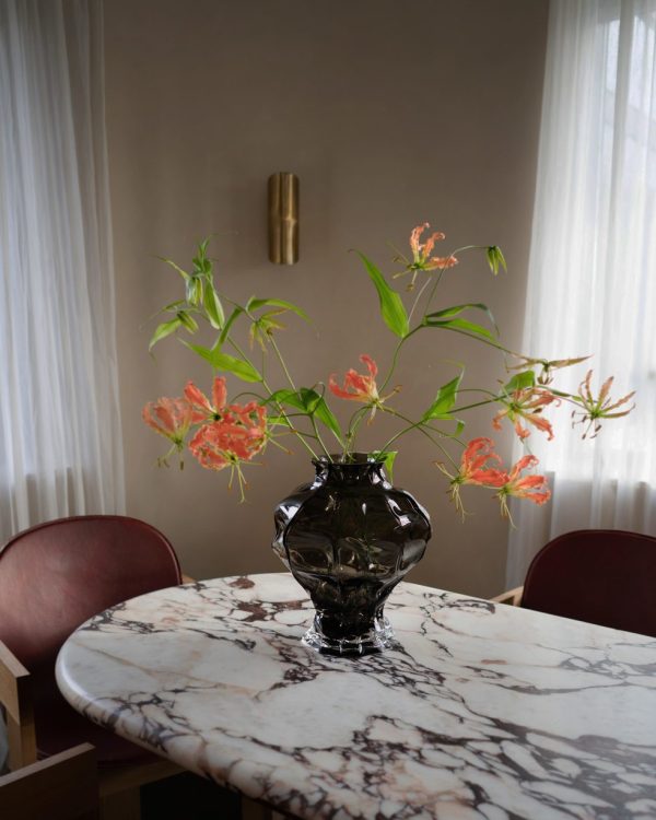 En mørk glasvase med levende orange blomster sidder på et marmorbord i et værelse med rene hvide gardiner og en subtil guldlampe på væggen. To røde designerstole flankerer bordet.