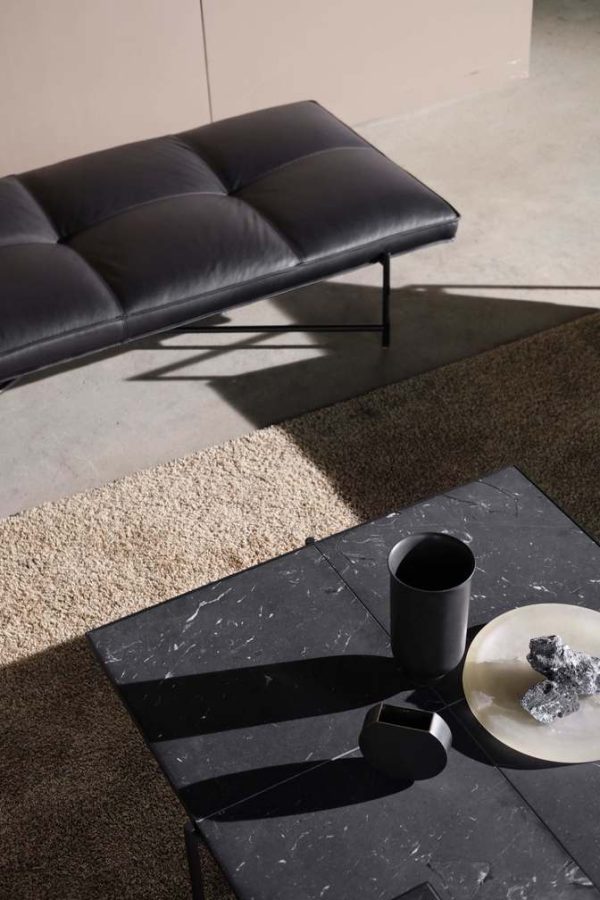En minimalistisk stue med et sofabord i sort marmor fra TomorrowsDesign med et krus og en tallerken med en croissant. En sort lædersofa og et beige tæppe er synlige, med sollys, der kaster skygger på scenen.