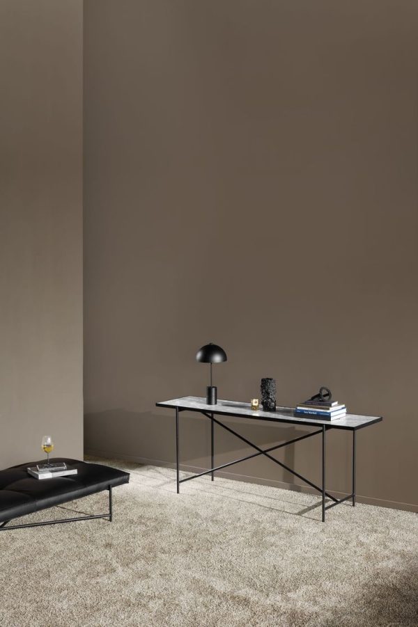 Et minimalistisk kontorsetup med et lækkert sort skrivebord, hvorpå der sidder en lille sort lampe, bøger og pynteting. Et matchende lavt bord med glasoverflade er i nærheden, alt sammen mod en almindelig, taupe væg, med interiørdesigndetaljer.