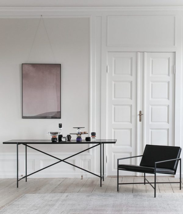 Minimalistisk interiør med et elegant sort skrivebord med forskellige småting ovenpå, en sort stol og et stort abstrakt maleri hængende på en hvid panelvæg. Lyse trægulve komplementerer den rene, rummelige æstetik.