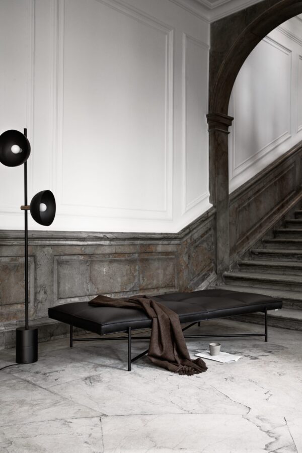 Et minimalistisk værelse med en lækker sort daybed og en moderne lampe ved siden af, placeret i et elegant rum med hvide panelvægge og en forvitret stentrappe, kurateret af TomorrowsDesign.