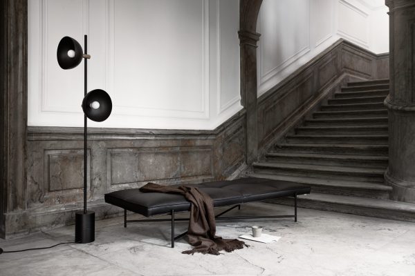 En moderne sort daybed med et brunt kast og en bog ved siden af, sat op mod en grå marmorvæg og trappe, med en stilfuld sort gulvlampe med to runde skærme. Dette interiør er perfekt til afslapning.
