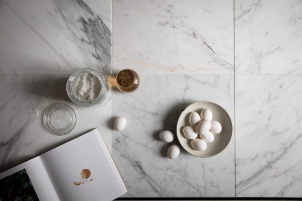 Set ovenfra og ned af en marmorbordplade med bageingredienser: en skål med æg, mel i en krukke, en krukke med brun farin og en åben kogebog med en synlig udtværing fra TomorrowsDesign.