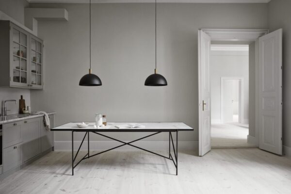 Et minimalistisk køkken med grå vægge og hvide gulve omfatter et bord i industriel stil, hængende sorte lamper og indbyggede hylder designet af TomorrowsDesign. En døråbning fører ind til et andet rum.