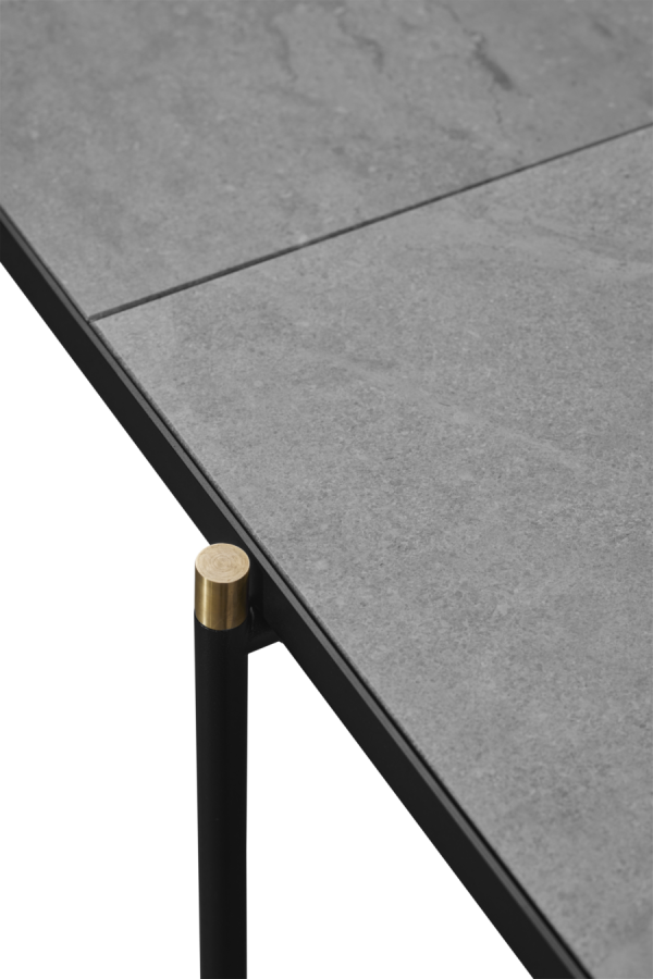 Nærbillede af et moderne bordhjørne med en slank, grå stenoverflade med synlige årer, understøttet af et slankt sort designmetalben med guldspids. Baggrunden er en almindelig, mørk gradient.