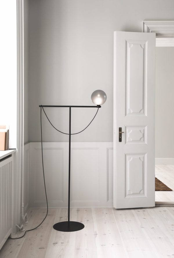 En åben hvid dør, der fører til et andet rum, med en slank, sort gulvlampe med en rund pære placeret i nærheden af den. Rummet har hvide vægge og lyst trægulv, der fremviser minimalistisk designmøbler.