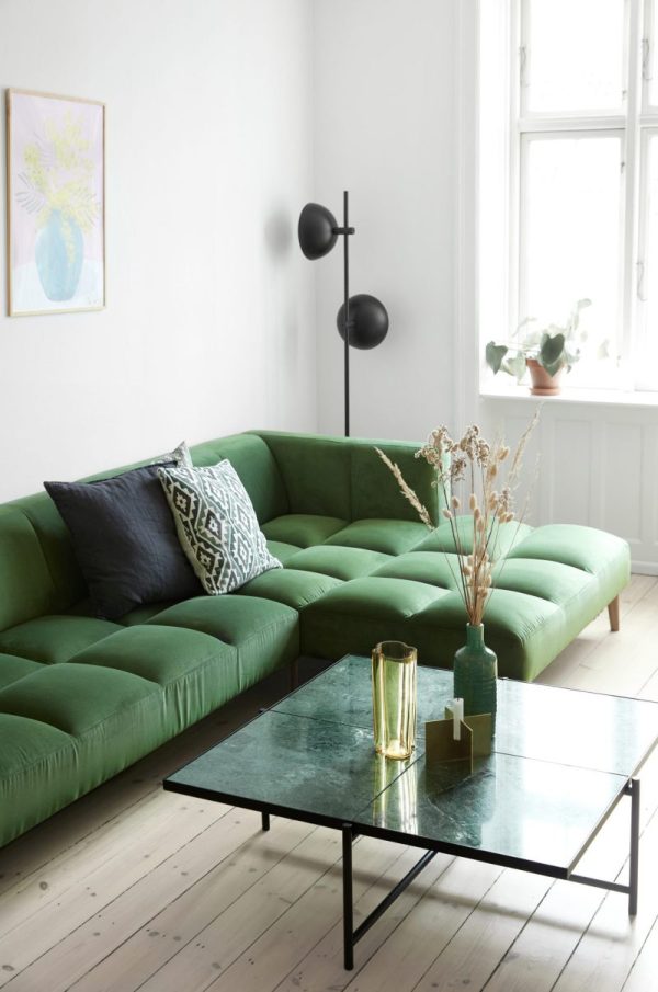 En lys, minimalistisk stue med en stor smaragdgrøn sektionssofa med mønstrede hynder, et elegant sort sofabord, en gulvlampe og dekorative planter ved et vindue. Lyse trægulve og en bleg væg fremhæver den luftige stemning. Indstillingen fuldendes af elegante designmøbler, der tilføjer