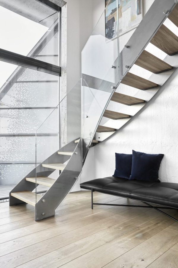 Et moderne interiør fra TomorrowsDesign med en træ- og metaltrappe med glasrækværk ved siden af en sort lædersofa med to mørkeblå puder. Rammerne omfatter en hvid væg og et vindue med mønstret matteret glas.