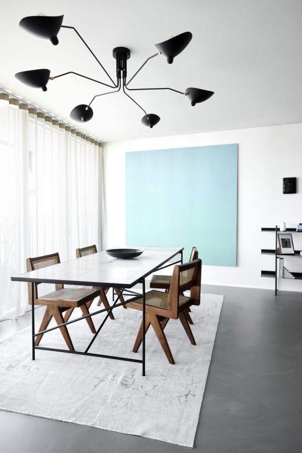 En moderne spisestue med et sort bord med fire stole, et stort abstrakt blåt maleri på væggen og en unik TomorrowsDesign loftslampe. Rummet har hvide vægge og et gråt gulv, fremhævet af et hvidt tæppe.