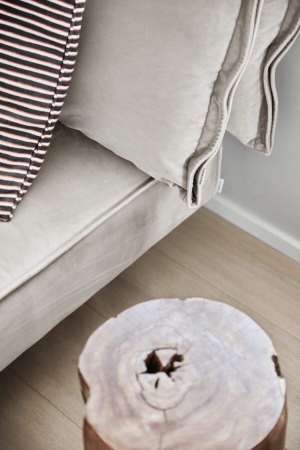 Et billede, der viser et nærbillede af en sofa med en beige fløjlspude, en stribet pude og en grå pude. I forgrunden fungerer et poleret tværsnit af en træstamme som et rustikt interiørbord.