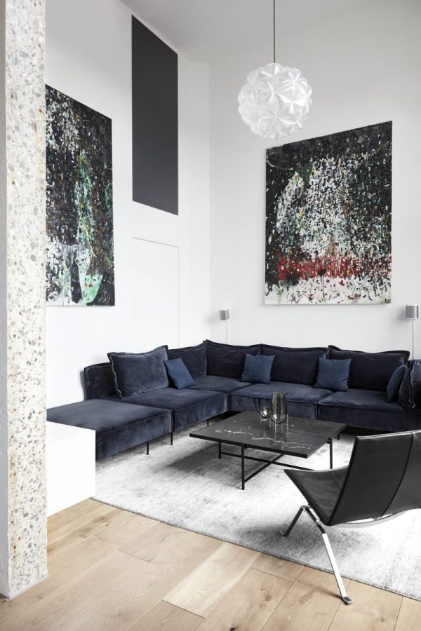 En moderne stue med en marineblå sektionssofa, et sofabord i sort glas, hvide vægge og abstrakte malerier. Der er også en unik hvid pendel og en minimalistisk TomorrowsDesign stol.