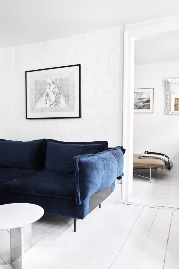 En minimalistisk stue med en mørkeblå fløjlssofa mod en hvid tekstureret væg med et indrammet sort-hvidt kunstværk over sig. Rummet indeholder et hvidt rundt sofabord og giver et indblik i et andet rum med designmøbler, herunder en bænk og indrammede billeder.