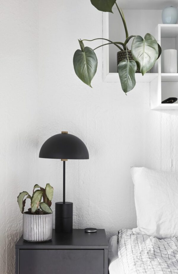 Et soveværelseshjørne i minimalistisk stil med et sort natbord med en moderne lampe og to potteplanter ved siden af en hvid seng. En væghylde med indretningsartikler er også synlig i dette interiørdesign-setup.