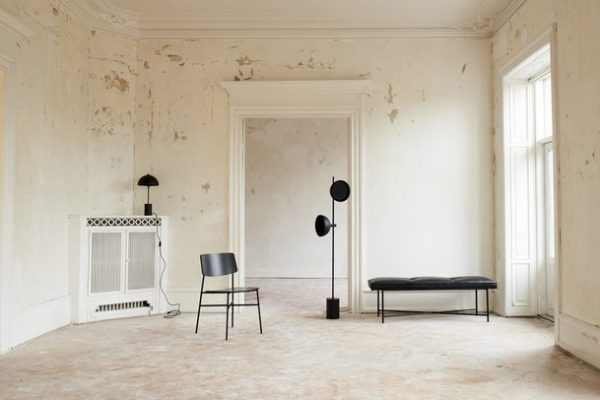 Et minimalistisk værelse med afskallet hvid maling, med designmøbler som en sort stol, gulvlampe og en bænk nær store vinduer, der fremhæver rummets vintage-æstetik og rigelige naturlige lys.
