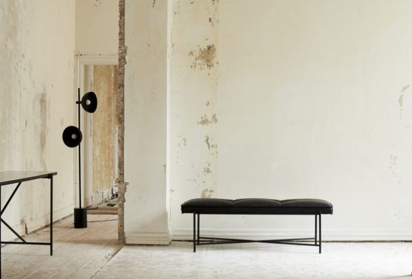 Indvendigt billede af et sparsomt rum med afskallede hvide vægge, med en sort læderbænk på en metalramme designet til komfort og stil, et lille rundt bord og to vægmonterede sorte lamper.