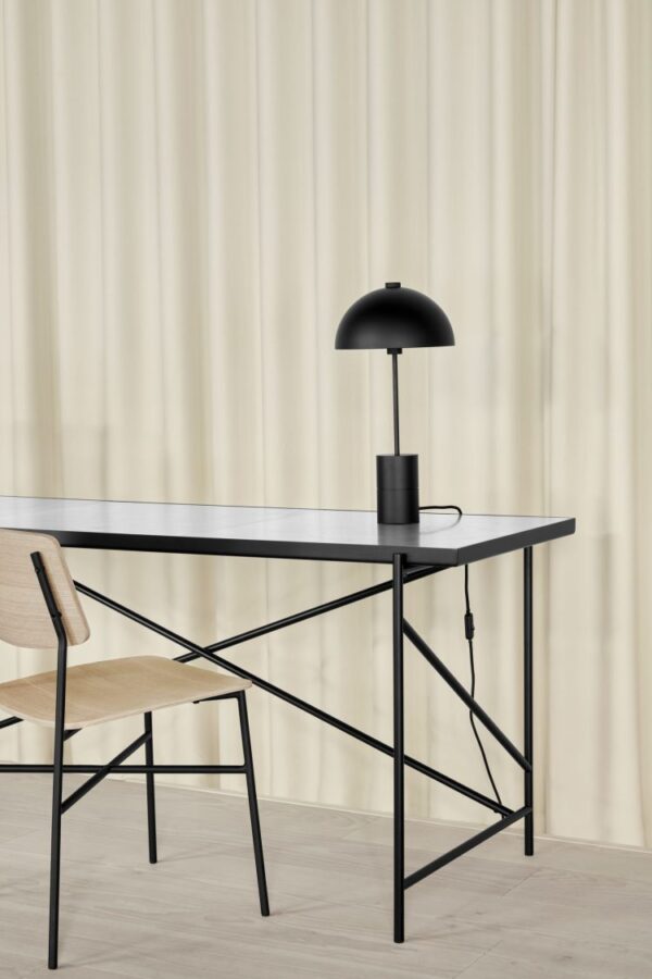Et moderne arbejdsområde med et elegant sort skrivebord med en x-formet metalramme, ledsaget af en lys træstol fra TomorrowsDesign og en sort kuppelformet skrivebordslampe sat mod en draperet beige gardinbaggrund.