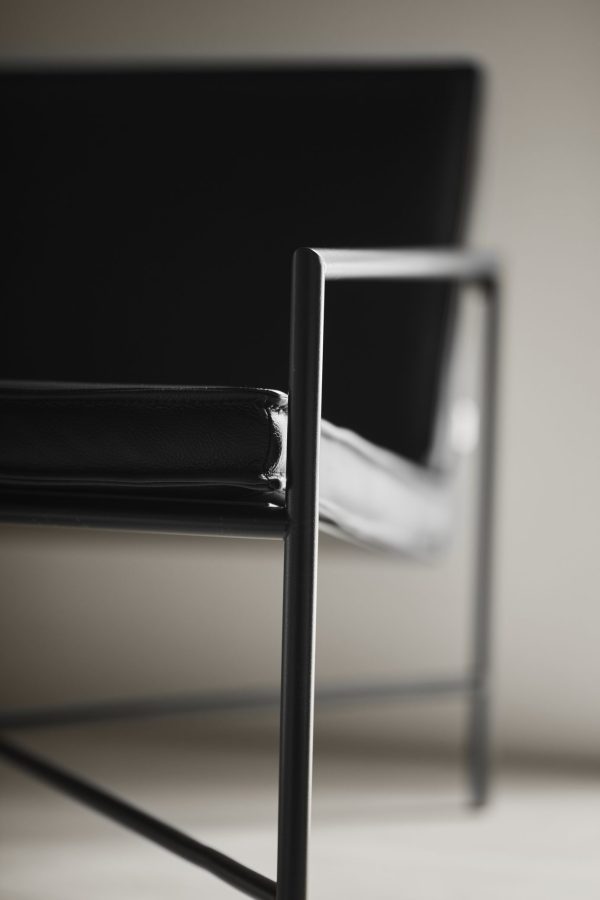 Nærbillede af en moderne stol med sort læderbetræk og en slank, tynd metalramme, der understreger teksturerne og det minimalistiske TomorrowsDesign. Baggrunden har en lys cremeoverflade, der ikke er i fokus.