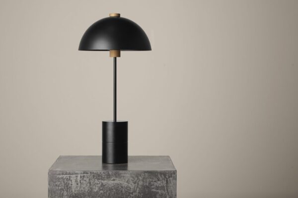 En moderne sort bordlampe med en afrundet skærm og cylindrisk base, stående på en tekstureret grå blok mod en neutral baggrund, inkarnerer essensen af TomorrowsDesign.