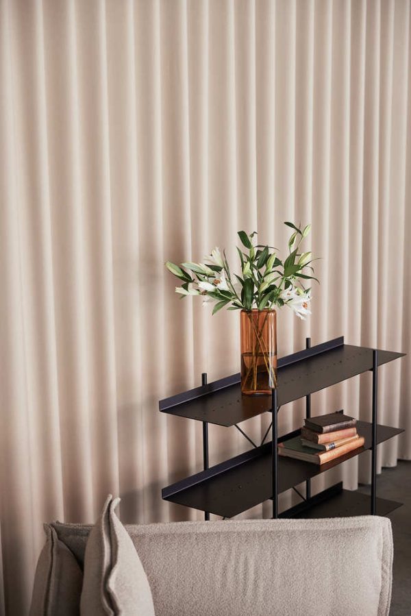 Et moderne design mørkt reol med bøger og en vase med hvide liljer mod en plisseret beige vægbaggrund, delvist skjult af en grå sofa.
