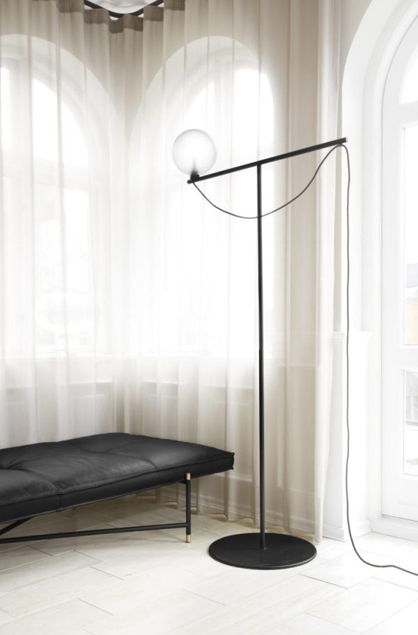 En moderne gulvlampe med et cirkulært lys og en buet sort ramme står ved siden af en læder daybed i et rum med gennemsigtige gardiner og sildebensgulve, som viser innovativt design.