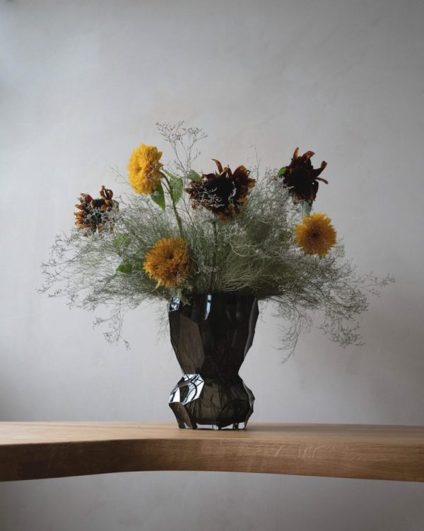 A HEIN STUDIO - Reflection Vase - New Smoke på en træoverflade, indeholdende et arrangement af luftige gule og mørkebrune blomster spækket med sart grønt løv mod en grå baggrund.