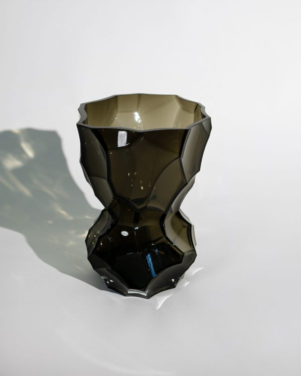 A HEIN STUDIO - Reflection Vase - New Smoke med en blank finish står på en reflekterende overflade og kaster en skygge. dens facetterede design skaber et kantet og moderne look.