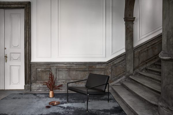 En minimalistisk lobby med en sort metalstol, designet af TomorrowsDesign, en terracottagryde med bregner og en lille rund bakke på et gråt tæppe. Baggrunden omfatter en hvid dør, grå marmortrapper og klassiske vægpaneler.
