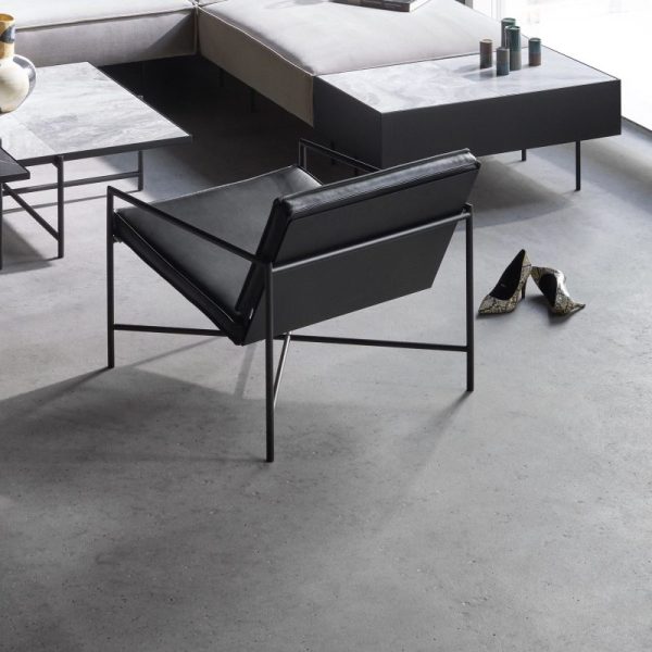 En minimalistisk stue med en stol med sort metalstel, parrede sofaer, et betongulv og spredte genstande, herunder et par høje hæle og bøger. Rummet afspejler TomorrowsDesign-æstetik med sine slanke designelementer.
