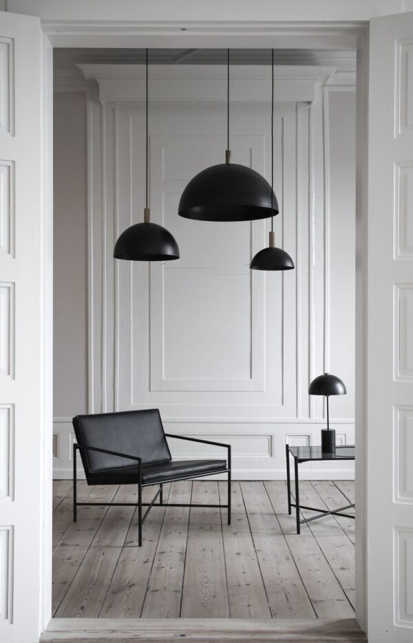 Et minimalistisk værelse med hvide vægge og trægulve, med en sort stol, et bord og tre hængende pendler. To døre er delvist synlige på siderne. Designet fremhæver interiørets enkelhed og elegance.
