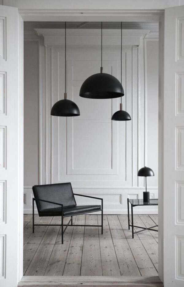 Et minimalistisk og elegant interiør med hvide panelvægge og trægulve, med to store sorte pendellamper, der hænger fra loftet, en elegant sort loungestol og en lille sort bordlampe på gulvet.