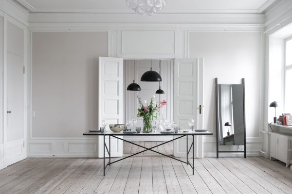 En minimalistisk spisestue med et slankt sort bordsæt til to, med et lyserødt blomstret midtpunkt. Hvide panelvægge, trægulve og *designmøbler* skaber et elegant rum.