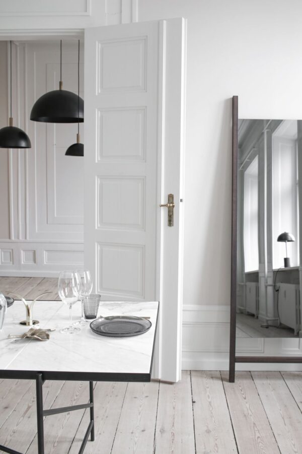 En moderne spisestue med en hvid dør på klem, der afspejler et udsnit af rummet med designmøbler inklusiv et spisebord i træ, glasvarer og en dekorativ tallerken, sat op mod hvide panelvægge og trægulve.