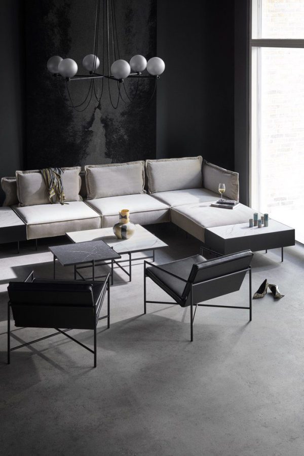 Moderne stue med minimalistisk design med en modulær grå sofa, HANDVÄRK FURNITURE - Sofabord 90 sofabord og stol med sort metalstel og en lysekrone. betongulv og stort vindue, med et par sko på gulvet.